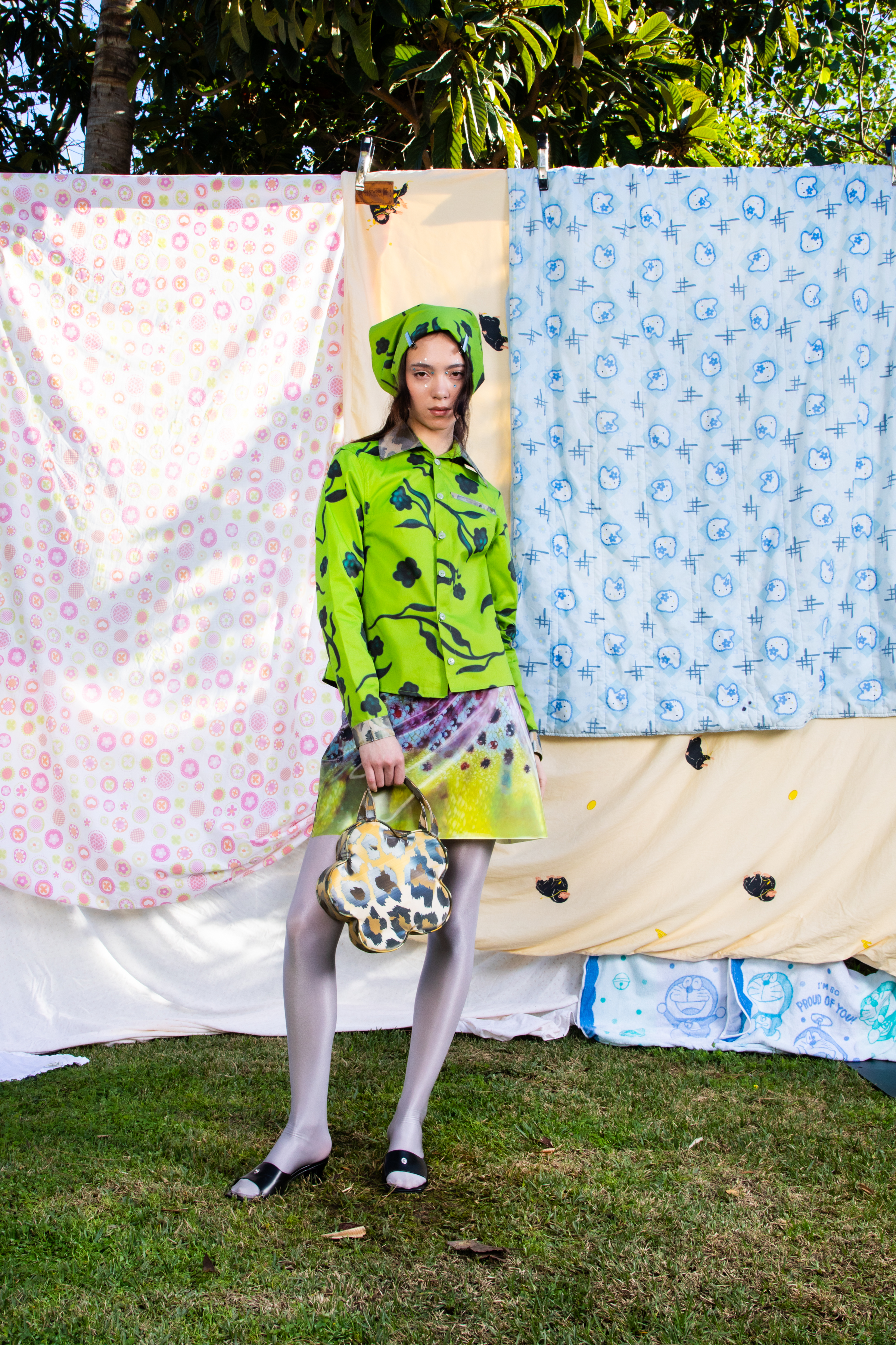 Full body image of a female model in a matching green patterned set with head scarf and matching flower shaped bag against a backdrop of patterned quilts.
